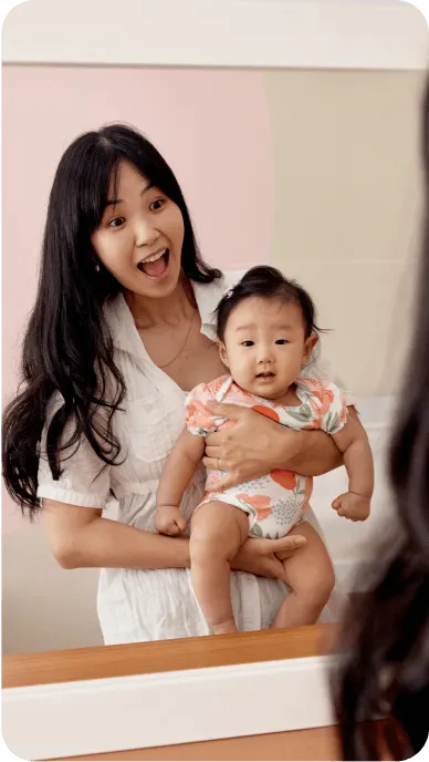 Woman smiles into mirror holding baby wearing floral jumpsuit