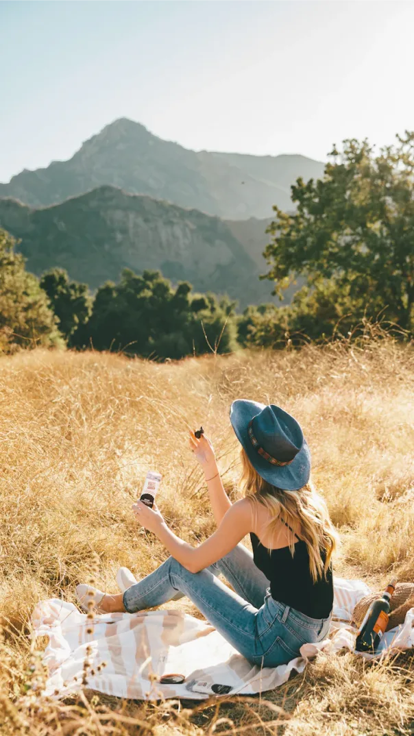 Woman with blue hat sits on blanket in a field facing mountains eating a packaged snack bar
