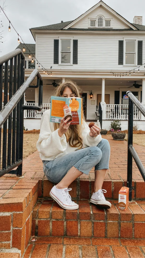 Woman sits on stairs outside of a large house reading a book and eating a packaged snack bar